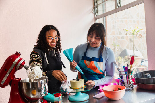 Happy woman helping friend decorating cake using icing bag at home