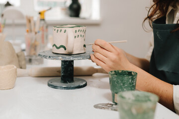 Fototapeta premium Painter is drawing and adding unique designs to the earthenware. Artist is decorating and painting handmade ceramic clay cup in the creative workshop.