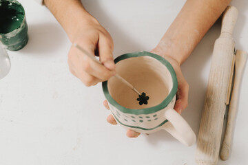 Female artist is painting earthenware with paintbrush and water colors in the workshop. Painter is drawing and making a colorful decoration of a clay ware.