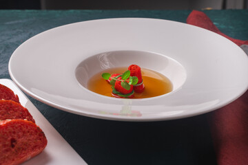 Consomme or broth with red omelette rolls served in a white plate with a red sesame bun on a dark green background with a napkin and spoon.