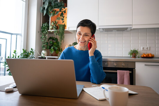 Smiling mature woman speaking on smartphone, working on laptop at home. Business call at remote work, freelancing, self employed occupation, managing from home office, professional multitasking