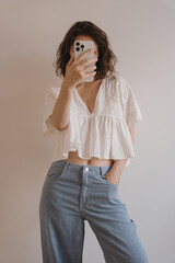 Woman in White Top and Denim Taking Mirror Selfie with Smartphone in Simple Room. Minimalist...