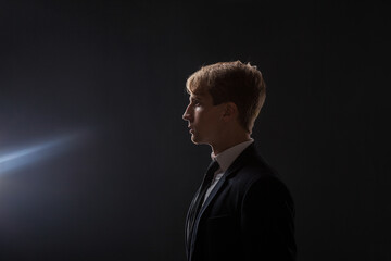 In a dark studio, a man in a black suit and white shirt stands in profile. The contrast between his attire and the shadowy background creates an air of mystery and professionalism. © Ulia Koltyrina