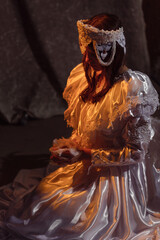 Beautiful young brunette dressed as an angel kneels in a dimly lit room, draped in an ornate white...