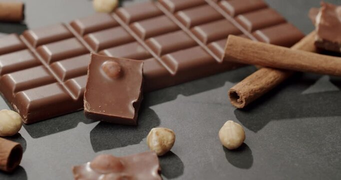 Close-up of a milk chocolate bar with hazelnuts, cinnamon sticks, and chocolate pieces on a dark surface, evoking a sense of indulgence and sweetness. Delicious Milk Chocolate Bar