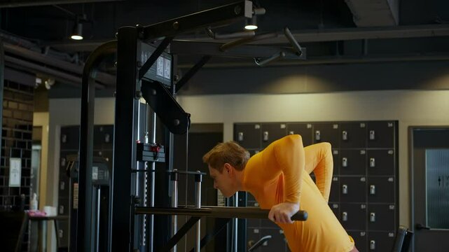 A man wearing a bright orange long-sleeve top and burgundy pants performs a tricep dip exercise using parallel bars in a modern gym facility with dark lockers visible in the background