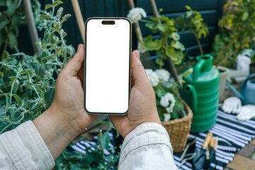 Hands holding a smartphone with a blank screen in a garden setting. A green watering can, potted plant, gardening tools, gloves, and striped cloth are in the background