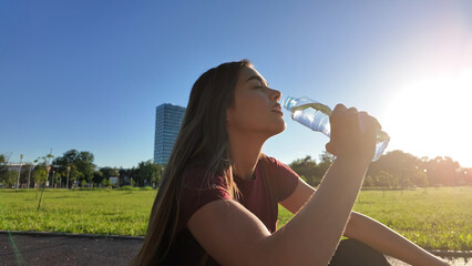 Young exhausted woman drinking water after jogging and exercise in urban park.