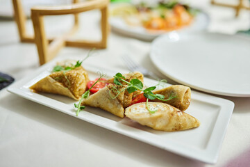 A plate with a variety of dishes, including a roll of pancakes stuffed with tomatoes on top