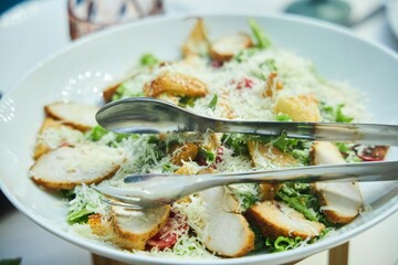 On a white plate there is a salad with chicken, a fork and a knife. The salad is decorated with cheese and lettuce leaves. Caesar salad with chicken