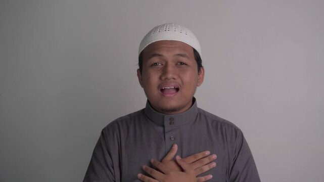 Southeast Asian Muslim man wearing grey robe makes greeting gesture, then chats amiably with people around him