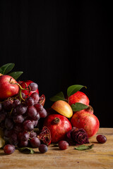 Still life with red apples, pomegranates and grapes on wooden table, black background, vertical with copy space