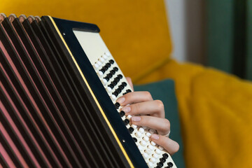 Close-up of female musician's hand playing a traditional accordion, with a blurred yellow sofa in the background, creating a vibrant and musical atmosphere