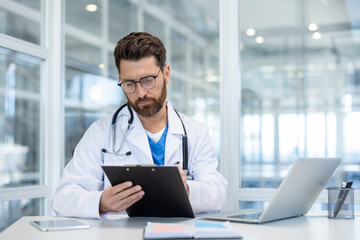 Concentrated doctor with beard and glasses is sitting at a desk and carefully reviewing patient information on a clipboard in his office