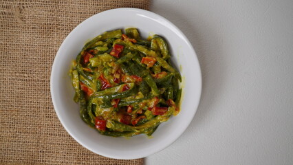 A portion of Balinese jukut bejek mixed with sambal embe made from long beans is served in a white container placed on a burlap base with a white background (isolated white).