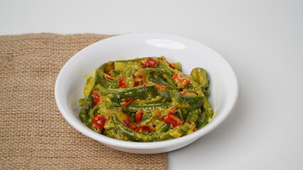 A portion of Balinese jukut bejek mixed with sambal embe made from long beans is served in a white container placed on a burlap base with a white background (isolated white).