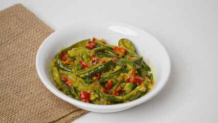 A portion of Balinese jukut bejek mixed with sambal embe made from long beans is served in a white container placed on a burlap base with a white background (isolated white).