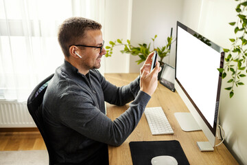 Naklejka premium Side view of cheerful young man sitting at home office typing a message and smiling at his phone.