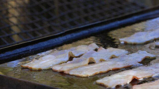 Close up: Strips of bacon fry on large griddle, strong diagonal line