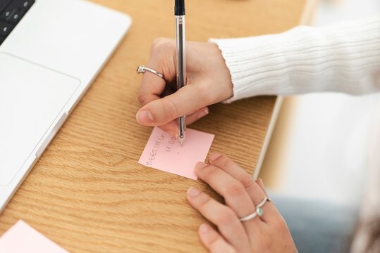 A close-up of a woman's hands writing a meeting reminder on a pink sticky note with a pen, placed on a wooden desk near a laptop in a workspace.