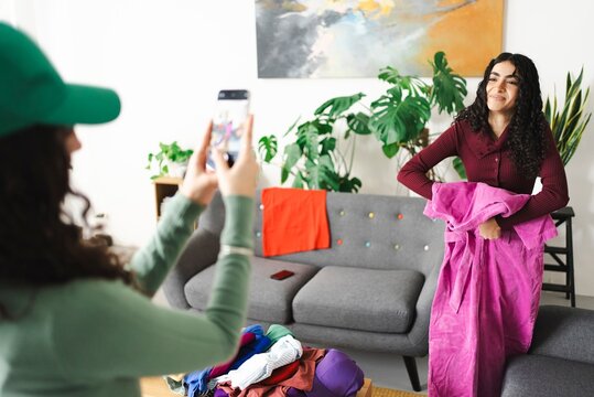 A young woman with curly hair is posing while holding a bright pink outfit as her friend captures the moment on a smartphone, in a stylish and modern boutique setting.