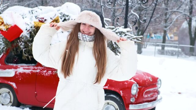 A beautiful young woman in a winter hat with earflaps next to a retro red car decorated for Christmas. A snowy winter day, christmas tree on car roof.