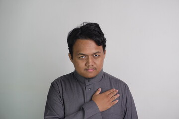Obraz premium Southeast Asian Muslim man wearing grey robe placing his hands on his chest crossed, with expressive expression
