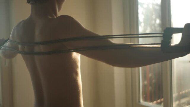 Athletically attractive teenager doing exercise using elastic band with handles for grip. Young guy stretches extended expander behind his back. Exercise for strengthening muscles. Sports for youth