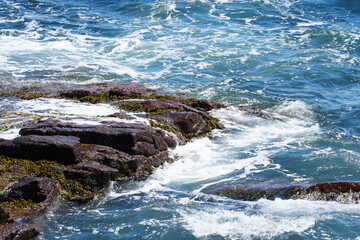 Powerful ocean waves crash against a rugged rocky shore, creating white foam and splashes. The dynamic scene captures the energy of the sea, perfect for travel, nature, and adventure themes.