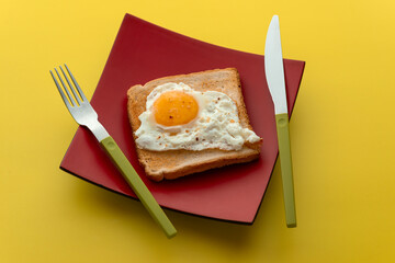 Scrambled eggs on toaster fried bread on quadrangular red plate and a fork with a knife with green handles on  a yellow table