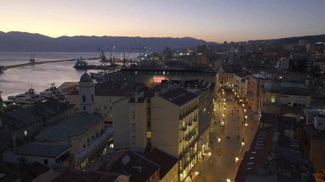 A nighttime aerial view of Korzo in Rijeka, Croatia, with city lights illuminating historic streets, the distant sea reflecting lights, and the 3. Maj shipyard cranes towering over the horizon.