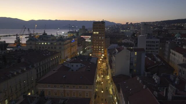 Aerial night view of Rijeka&rsquo;s Korzo, where glowing city streets meet the dark sea, with the illuminated cranes of the 3. Maj shipyard standing tall in the distance.