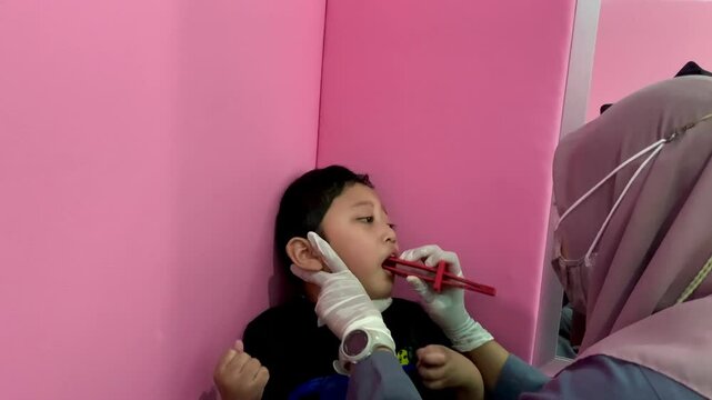 A child with cerebral palsy is undergoing speech therapy with a therapist at a child growth and development clinic, leaning against a wall covered in pink foam for support and guidance.