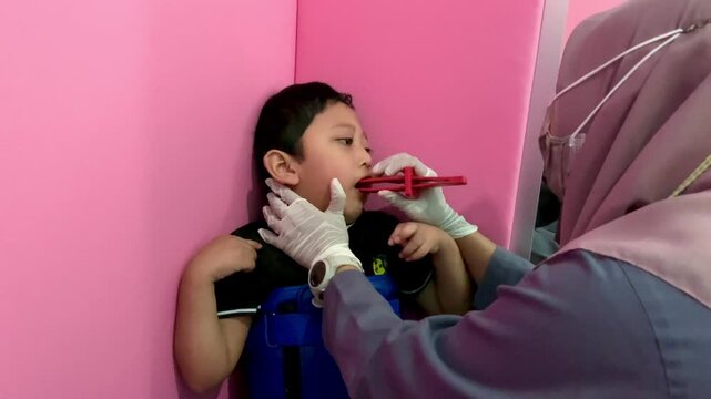 A child with cerebral palsy is undergoing speech therapy with a therapist at a child growth and development clinic, leaning against a wall covered in pink foam for support and guidance.