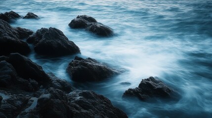 Obraz premium Soft waves caress dark, rugged rocks on a quiet shore, capturing the harmony and gentle persistence of nature under a moody sky.