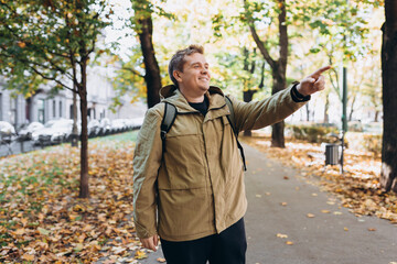 Obraz premium Blonde young 30s man wearing backpack pointing something on city autumn park street. Urban lifestyle, travel concept. High quality photo