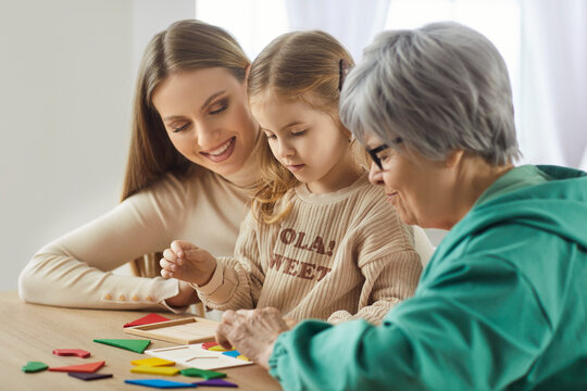 Side view of three generations of women enjoying time together, playing with colorful educational shapes. Smiling grandmother, mother and granddaughter stacking jigsaw pieces and having fun at home.