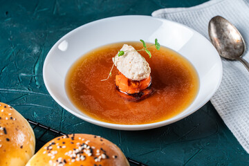 Soup or consomme, thick meat broth with carrots, chicken dumplings and microgreens, served in a white plate on a dark green background with sesame seed buns, napkin and spoon. View from above