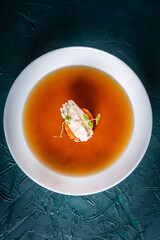 Soup or consomme, thick meat broth with carrots, chicken dumplings and microgreens, served in a white plate on a dark green background with sesame seed buns, napkin and spoon. View from above