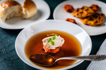 Soup or consomme, thick meat broth with carrots, chicken dumplings and microgreens, served in a white plate on a dark green background with spoon. View from above. Space for text. Flat lay.