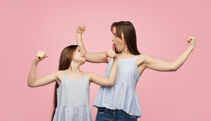 Happy adult woman with little girl wearing similar clothes and showing biceps happily posing on pink background