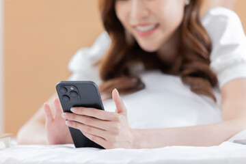 Young asian woman is looking on smartphone screen while comfortably  lying on her bed in day time