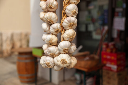 A close-up of a braid of garlic bulbs hanging outside a traditional market. The off-white bulbs with dried stems are woven together for easy storage and preservation
