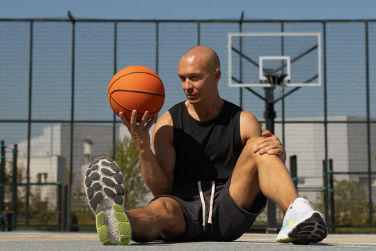 Basketball player resting with orange basketball ball and looking at ball on sports field. Game interval. Outdoors.
