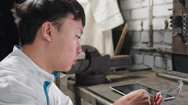 Young lab technician working with veiner caliper adjusting measurements, tapping on tablet, returning to adjust caliper on boris placed on table, background includes mechanical tools