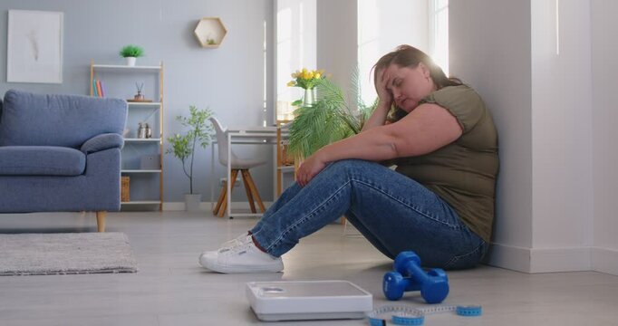 Upset fat plus-size woman looking sadly at gym equipment at home, showing reluctance toward workout routines. He feeling problems and challenges of fitness and training motivation.