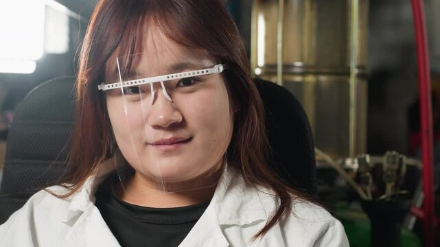 Student wearing protective face shield blinks eyes with warm smile in mechanical workshop, background features industrial tools, bright lighting, and machinery