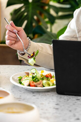 A close-up of a hand holding a fork with a fresh salad, while another hand rests on a laptop. The setting suggests a working lunch, combining healthy eating with productivity.