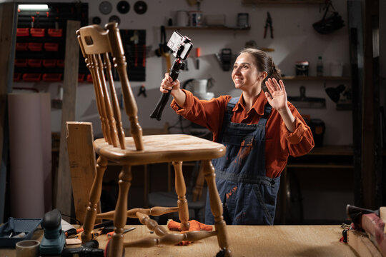 Female carpenter holding camera on tripod while recording diy video blog, she restoring old furniture in her workshop