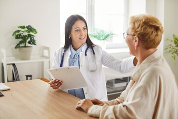 Obraz premium Young woman doctor in white lab coat holding clipboard, consulting with older female patient in bright medical office. Friendly cardiologist providing support and discussing health recommendations.